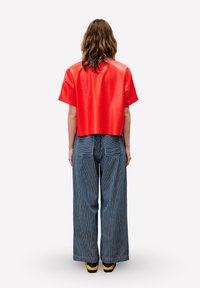 Rode cropped glanzende top gecombineerd met wijde, blauw-wit gestreepte broek. Model draagt bruine schoenen met gele accenten.