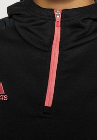 Pull noir avec capuche et fermeture éclair rose. Dispose d'un petit logo brodé rouge sur la poitrine gauche et d'un tissu texturé.