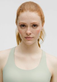 Top atlético de color verde claro con una textura suave, tirantes anchos y escote redondeado. La modelo tiene el cabello rojo y pecas.
