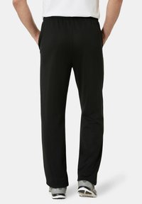 Mann steht mit den Händen in den Taschen, trägt schwarze Hose mit elastischem Bund und graue Sneakers, von hinten vor einem weißen Hintergrund dargestellt.