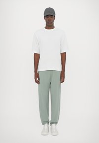 Bijela majica s kratkim rukavima u kombinaciji sa svijetlozelenim trenirkama i bijelim tenisicama, uz dodatak sive kape. Jednostavan dizajn i meki materijali.