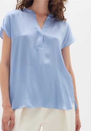 Blusa azzurra chiara a maniche corte in tessuto liscio con scollatura a V e spacchetti laterali, abbinata a pantaloni color crema.