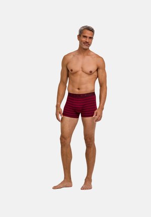 Fit Mann mit kurzen grauen Haaren steht barfuß da und trägt rote und dunkelrote gestreifte Boxershorts vor einem schlichten hellen Hintergrund.