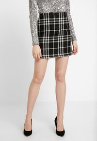 Falda mini de cuadros en blanco y negro, hecha de una tela texturizada. Se lleva con una blusa de mangas largas brillante en plata y tacones altos negros.