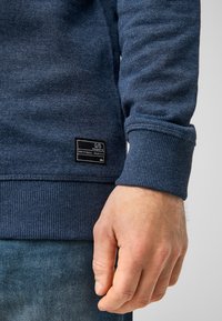 Sudadera azul marino con puños acanalados, tejido texturizado y una etiqueta en el lado inferior. Corte ajustado con ligero estiramiento y superficie suave.