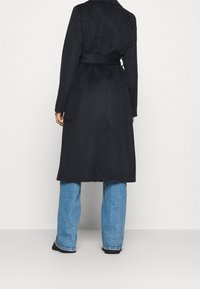 Lang sort uldfrakke med bælte i taljen, dobbeltradet design og to sidelommer, kombineret med lyseblå vidde bukser.