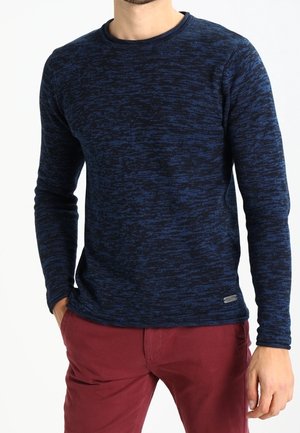 Uomo che indossa un maglione a maniche lunghe blu scuro con motivo e pantaloni rossi, con una mano in tasca contro uno sfondo bianco.