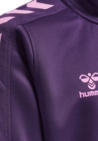 Paarse sportjasje van glad, rekbaar materiaal. Heeft roze diagonale strepen en een "hummel" logo op de borst.