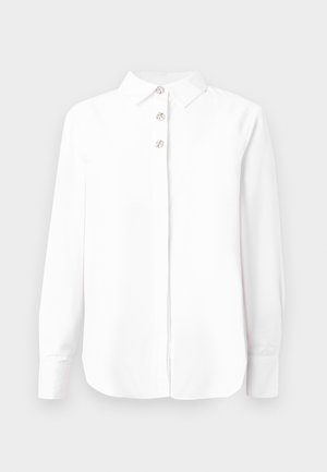 Bijela košulja s dugim rukavima, klasičnog ovratnika i četiri dekorativna gumba sprijeda. Glatka tkanina s laganim sjajem.