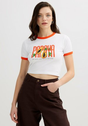 Joven mujer de cabello castaño lleva una camiseta corta blanca con ribete naranja y gráfico de papaya, combinada con pantalones marrones de cintura alta, mano en el bolsillo.