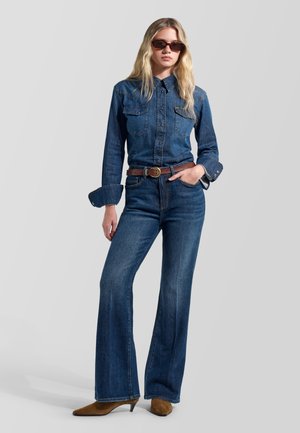 Giovane donna che indossa una camicia di jeans blu scuro e jeans con cintura marrone, scarpe marroni con tacco e occhiali da sole scuri, in piedi con una mano in tasca.