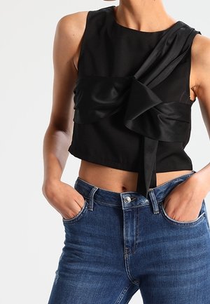 Kvinde iført sort, ærmeløs cropped top med stor sløjfedetalje og blå denimjeans, hænderne puttet i forlommerne.