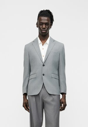 Man draagt een lichtgrijs colbert met twee knopen en bijpassende broek over een wit overhemd met kraag, staand tegen een effen achtergrond.