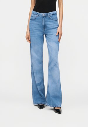 Person, die hellblaue, hochtaillierte Jeans mit weitem Bein und schwarze spitze Schuhe trägt, steht vor einem einfarbigen hellen Hintergrund.