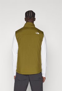 Olivgrön vadderad väst med hög krage, med en liten vit logotyp på vänster axel, bärs över en vit långärmad skjorta.