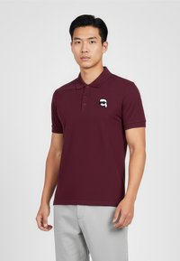 Burgundská polokošile vyrobená z hladké tkaniny, s límečkem a malým bílým logem na levé hrudi. Krátké rukávy a zapínání na knoflíky.