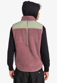 Fleece-Weste mit einem rosa, strukturierten Oberteil und einer Schulterpartie aus grünem, einfarbigem Stoff; mit hohem Kragen und Zugband auf der Rückseite.