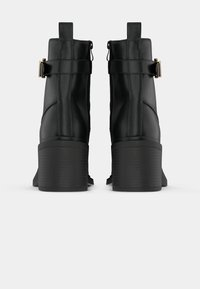 Paire de bottines noires à talons blocs avec des brides latérales munies de boucles métalliques, vues de dos sur fond blanc.
