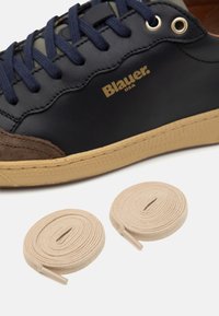 Baskets bleu marine et marron avec semelle beige, lacets bleu marine et deux lacets roulés beige sur fond blanc.