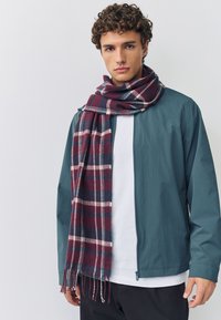 Sciarpa a quadri in bordeaux, blu navy e crema appoggiata su una giacca turchese. La sciarpa ha bordi sfrangiati. La modella posa su uno sfondo bianco.