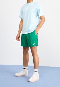 Světle modré tričko Nike, zelené šortky, bílé logo Nike. K tomu beige a růžové vyšší tenisky, doplněné černobílými pruhovanými ponožkami.
