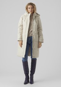 Manteau long matelassé de couleur beige clair avec une capuche, doté de fermetures à boutons et de poches latérales, associé à des bottes violettes brillantes et un jean bleu.