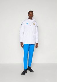 Balta sporta jaka ar kapuci un rāvējslēdzēju, ar zilām Adidas svītrām. Kombinēta ar zilām biksēm un melniem sporta apaviem. Redzams Real Madrid logotips.