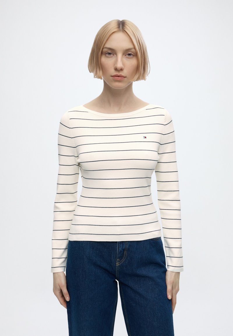 Femme aux cheveux blonds courts porte une chemise blanche à manches longues avec des rayures horizontales noires et un jean bleu, debout devant un fond clair uni.