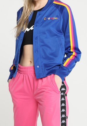 Mujer con chaqueta azul abotonada con franjas arcoíris y pantalones rosas con cinta lateral de marca en blanco y negro, con las manos en los bolsillos.