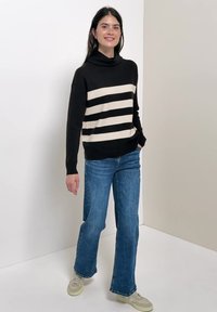 Femme portant un pull rayé noir et beige, un jean bleu et des baskets beiges, debout devant un fond blanc et beige uni.