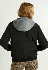 Chaqueta bomber acolchada negra con capucha gris. Presenta puños y dobladillo acanalados, mostrando una mezcla de texturas y sutiles patrones en forma de diamante.