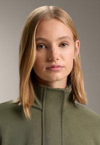 Olivovo zelený oblečenie s vysokým golierom, štruktúrovaným golierom, hladkou textúrou a minimalistickým dizajnom, s čistými líniami a bez viditeľného kovania.