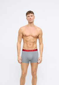 Muž bez trička stojí proti bielej stene a má na sebe šedé boxerky Nike s červeným pásom.