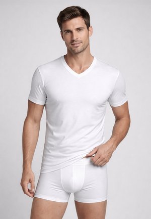 Uomo che indossa una T-shirt bianca a scollo a V e boxer bianchi, che tiene l'orlo della maglietta con una mano, su uno sfondo chiaro e uniforme.