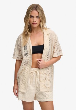 Mujer con cabello rubio largo lleva una camiseta beige de encaje de manga corta y pantalones cortos a juego con cordón encima de un top corto negro.