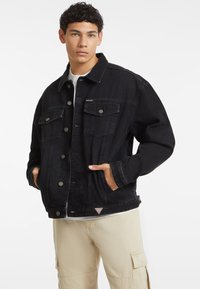 Giubbotto in denim nero con chiusura a bottoni, due tasche sul petto e tasche laterali. Il design include un colletto classico e un dettaglio logo discreto.