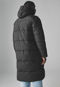 Sort puffy jakke med hætte, der har et quiltet mønster, lange ærmer og en knælængde design. Bæres over lyseblå jeans.