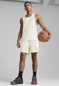 Débardeur et short de sport PUMA jaune clair, avec des accents roses. Baskets noires et rouges. Tenant un ballon de basket. Texture de tissu lisse.