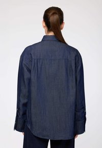Camicia di jeans blu scuro con maniche lunghe, taglio dritto e orlo leggermente curvato. Presenta polsini con bottoni e colletto classico.