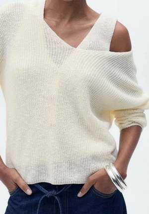 Femme portant un pull crème en maille ample avec une épaule découverte, assorti à un pantalon bleu foncé et des bracelets argentés au poignet.