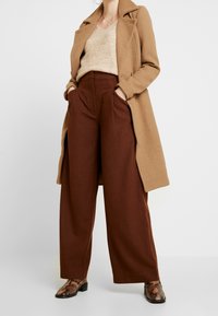 Vrouw in een beige jas, lichtbeige fuzzy trui, hoog getailleerde brede bruine broek en laarzen met slangenhuidpatroon.