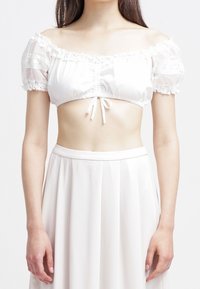 Donna che indossa un top corto bianco a spalle scoperte con pizzo e maniche voluminose, abbinato a una gonna plissettata a vita alta.