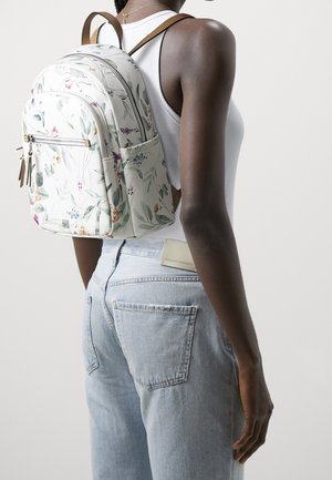 Person mit einem weißen Rucksack mit Blumenmuster, weißem ärmellosem Oberteil und hellblauen Jeans, von hinten und seitlich gezeigt.