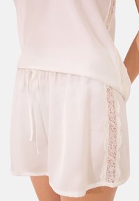 Ensemble de sommeil en satin blanc comprenant un haut à coupe ample et un short, avec une bordure en dentelle le long du côté des shorts et une taille élastique avec cordon de serrage.