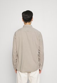 Beige pitkähihaispaita, jossa on nappikaulus, teksturoitu kangas ja yksittäinen takayökkä, napilliset kalvosimet ja rento istuvuus.