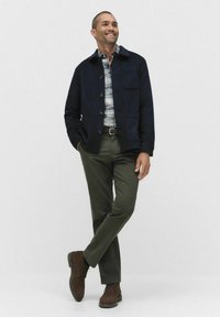 Man som bär en marinblå jacka, rutig skjorta, olivfärgade byxor och bruna skor, står med benen korsade och händerna i fickorna, leende.