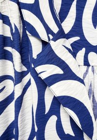 Tissu bleu et blanc avec de grands motifs abstraits de feuilles et des coutures visibles au niveau du col sur une surface de vêtement texturée.