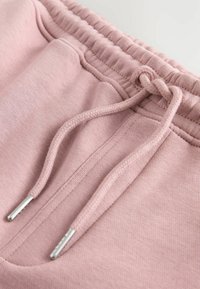 Pantalones de chándal rosas con cordón y puntas metálicas plateadas, cinturilla elástica y una tela suave y texturizada. Detalle del lazo y la tela mostrados.