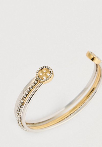 Atviras sidabro ir aukso apyrankė su kristalų akcentais ir apvaliais galais, turinti aukso emblemą, apsuptą mažų kristalų.