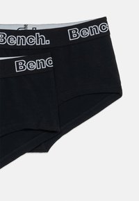 Svarte bomullsboksershorts med en glatt tekstur, med en hvit elastikk med merket "Bench.". To par som overlapper litt.